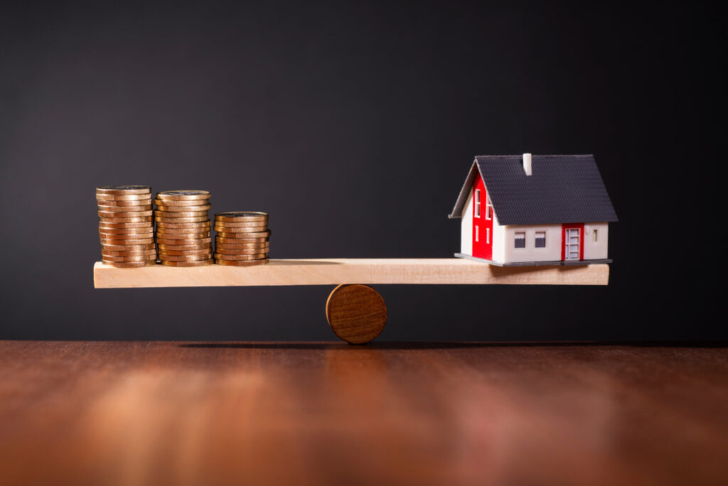 Wippe mit einem Haus auf der einen und Stapeln mit Münzen auf der anderen Seite, als Symbol der Baufinanzierung
